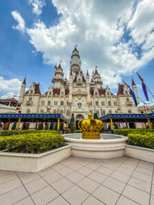 Photographie en perspective du château de Fort Fort Lointain à Universal Studios Singapore, avec une fontaine en forme de couronne dorée au premier plan, sous un ciel bleu nuageux.