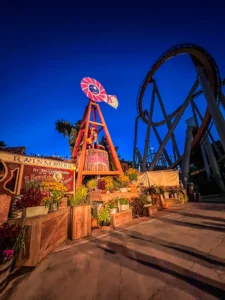 Un moulin à vent décoratif en bois et une charrette fleurie rétro "Rainmaker" illuminés à la tombée de la nuit, avec la structure imposante d'un grand huit en arrière-plan sous un ciel bleu foncé à Knott's Berry Farm.